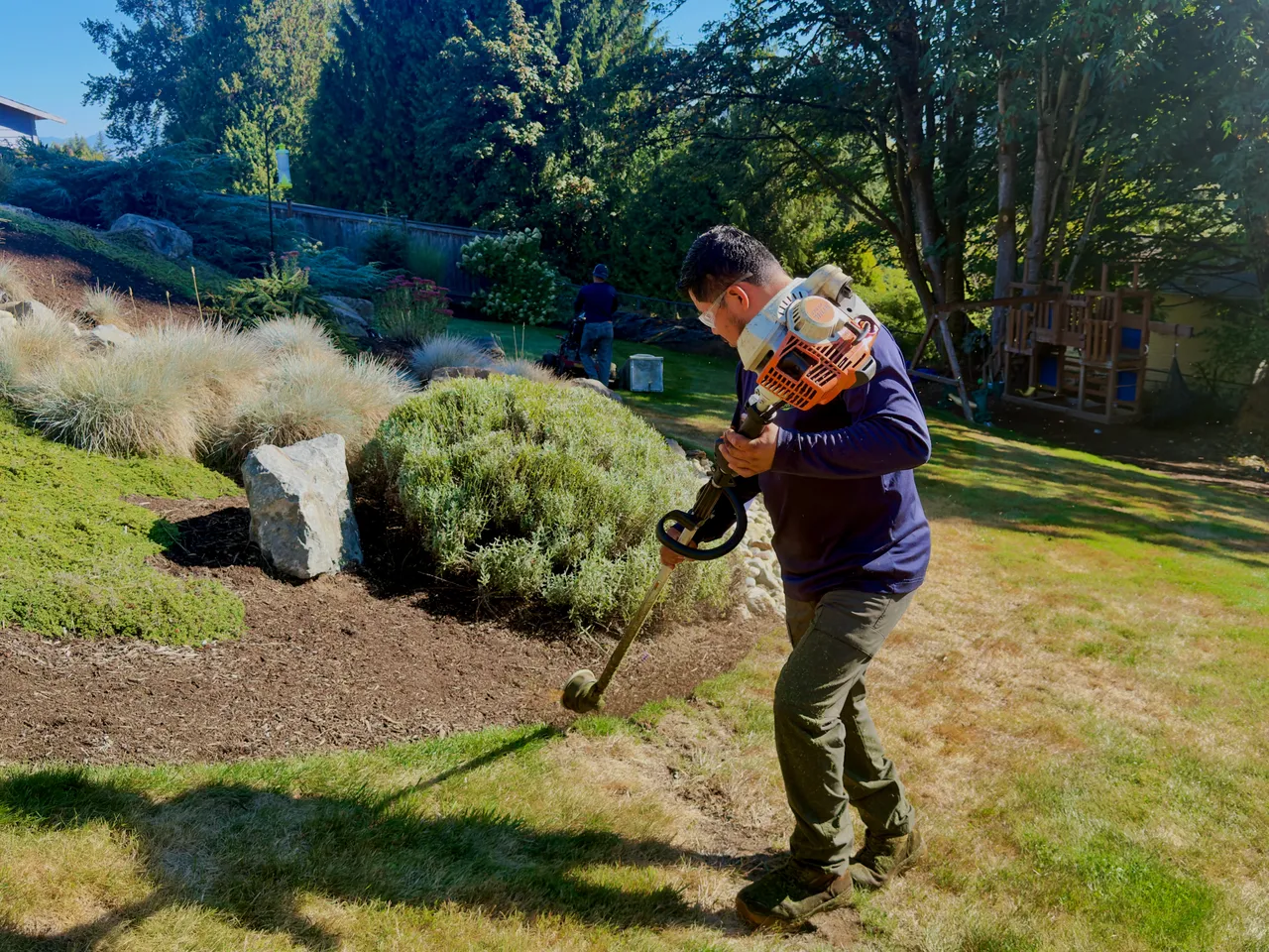 A crew member trims the bed edge on a sloped garden of grasses and boulders