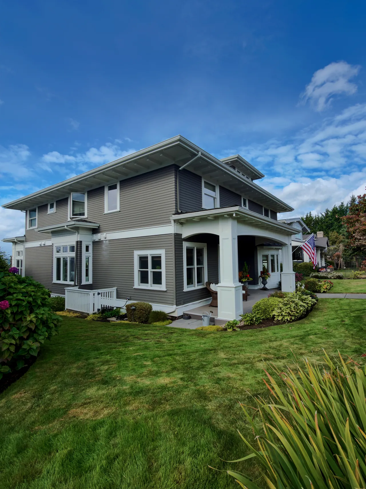 A grey foursquare house with a trimmed lawn and foundation beds, seen from the walk